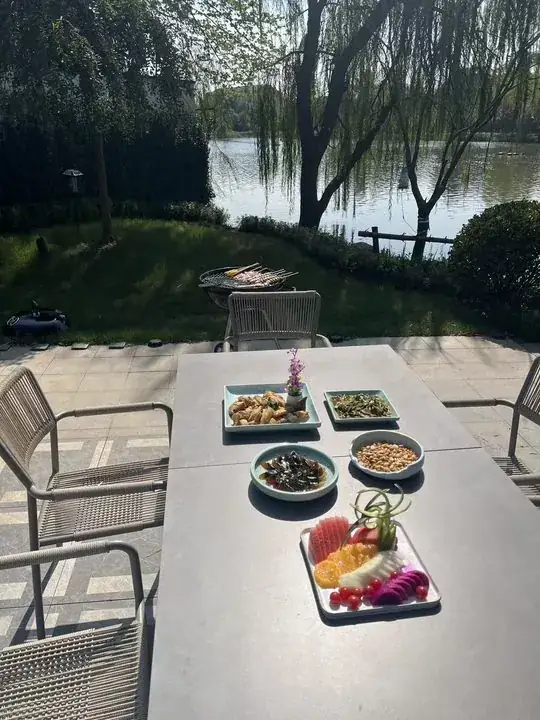 湖畔露台餐桌摆有水果和菜品,草地旁有烧烤架,背景是柳树与湖面